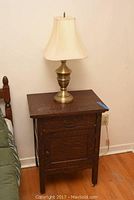 Full view of oak tiger wood side table with brass lamp on top, next to bed.