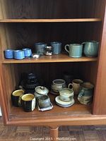 Shelf with green and blue glazed mugs, small bowls, a pitcher, and two metal tea strainers.