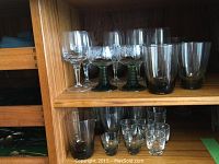 Shelf with assorted drinking glasses including wine glasses with clear and green stems, short tinted tumblers, and clear shot glasses on a wooden shelf.