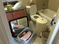 View of bathroom with toilet, bathtub including plastic baby bath seat, countertop with items including an untested DustBuster, decorative container, large mirror leaning against wall (damaged), beige clothes hamper with towels, plastic bucket, and mop visible.