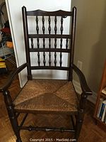 Full view of the solid wood chair with armrests and woven rush seat showing turned spindle backrest and arm supports.