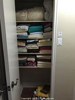 Open linen cupboard showing neatly folded stacks of white, teal, pastel, and patterned towels and sheets arranged on shelves.