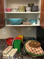 Wide view of cabinet with Pyrex bowls on middle shelf, assorted ceramic pots, plastic canisters, tins and bowls on counter below.