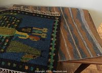 Two wool wall-hangings displayed on a wooden surface; one with a colorful figurative design on dark blue background, partially visible fringed edge.