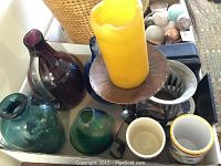 Various hand-formed glass vases and pitcher in purple, green, blue, teal colors along with a candle holder with yellow candle and ceramic cups shown inside a box.