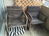 Two weathered wood deck chairs with horizontal slats and curved armrests, placed together on a covered balcony with lattice behind them. One chair cushion visible beneath seat.