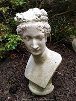 Front view of the cement bust statue showing the detailed facial features and hair curls, set in garden soil surrounded by greenery.
