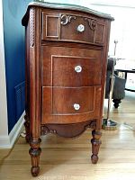 Front angled view of wooden end table showing three drawers, decorative scrollwork, glass knobs, and turned legs.