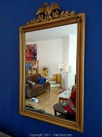 Front view of gilt framed mirror hung on a blue wall with visible eagle embellishment and decorative corner carvings reflecting a living room interior.
