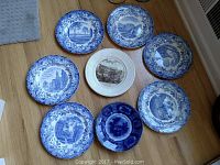 Seven plates laid out on a wooden floor including six blue and white Wedgwood plates and one different plate with a deep blue pattern.