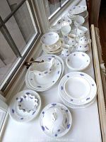 Wide view of the Adderleys Bone China pieces spread on a white surface near a window, showing plates, cups, pitchers, a gravy boat, and a covered dish with blue floral designs.