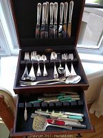 Wooden case with two drawers open showing silver flatware in top drawer and assorted knives in bottom drawer
