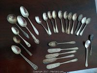 Full assortment of 21 silver spoons of mixed sizes and handle styles laid out on a dark surface, showing tarnish and varied wear.