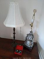 Two table lamps on wooden surface, one with cream fabric shade and dark wooden base, the other with an Asian figure base missing a shade, alongside a small open red case with stress balls