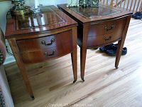 Set of two wood end tables with embossed leather tops, drawers, and caster wheels, placed on wooden floor with some wear visible on front of one table.