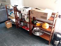 Three wooden folding shelves standing side by side, loaded with household kitchen items such as dishes, pots, bowls, and boxes. Shelves made of brown wood, showing smooth finish and foldable design.