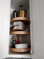Bakeware and kitchen items stored in a white corner cabinet on rotating shelves including springform pans stacked, insulated carafes, brass ice bucket on top shelf, and other small containers on bottom shelf.