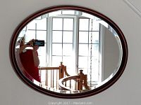 Oval beveled edge mirror with wooden frame hanging on a wall, showing reflection of window and staircase.