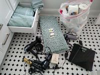 Photo showing the lot items on black and white tiled floor: two black hairdryers (one with diffuser), curling wand, digital scale, green bathmat with tags, green folded towels, and a plastic bag of bathroom paper products.