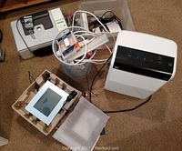 Photo of Dell printer, digital photo frame with stand, plastic bins containing electronic cables and accessories, and a white 35-pint dehumidifier unit on carpeted floor.