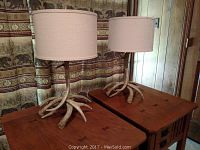 Pair of Pottery Barn synthetic antler base table lamps with beige drum shades on wooden side tables against patterned fabric backdrop.