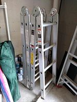 Folded aluminum convertible ladder leaning against a wall near paint cans and other items.