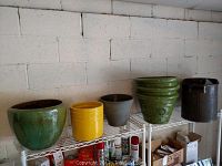 Side view of six garden pots including three green glazed ceramic pots, yellow, dark gray, and black plastic pots, and a large black plastic pot on the right, all arranged on a white wire shelf against a white wall.