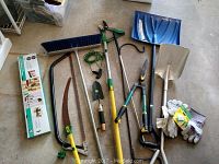Overview photo showing full lot of garden and cleaning tools including shovel, snow shovel, broom, saws, shears, and gloves laid on floor.