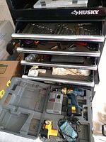 Husky tool chest drawer open, showing assorted wrenches, socket tools, scissors, and hand tools. Ryobi cordless drill kit with charger and battery in plastic carrying case below drawer.