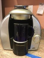 Front view of silver Tassimo espresso maker with a black mug placed on the base showing control knob and indicator lights.