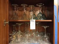 Glass stemware and tumblers stored on two shelves inside a wooden cabinet, including martini and wine glasses and tumblers with decorative gold leaf Japanese crane design.