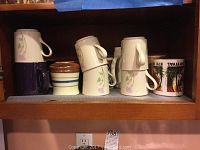Photo showing various coffee mugs including white ceramic mugs with purple floral design, a brown striped container, and a souvenir mug.