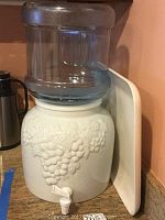 White ceramic water pitcher with grapes embossed on the sides, topped with a plastic water container and plastic tap spout at bottom. Also visible is a white ceramic tray leaning against the wall behind the pitcher.