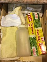 Drawer containing several kitchen dish towels in yellow, white, green, and blue, two rolls of Glad cling wrap, and a translucent plastic water bottle with rubber cap.