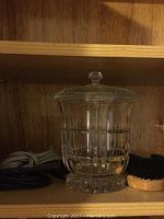 Clear glass vase with lid, showing etched decorative pattern, on wooden shelf.