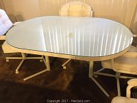 Elliptical white glass top kitchen table shown with chairs around it. The reflective glass surface shows vertical blinds reflection. The table has white metal legs and frame with some visible scuffs and chips.