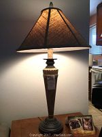 White antiqued resin lamp with dark brown triangular fabric shade on a table, lit to show the lamp details.
