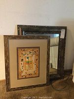 Photo of two framed items leaning against wall: one mirror with dark distressed frame and one framed abstract art print