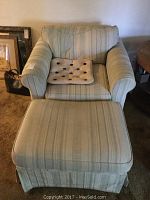 Front view of the overstuffed chair with a cushioned seat and rolled arms, upholstered in light-colored striped fabric, accompanied by matching ottoman with pleated skirt.