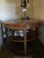 Front angled view of oak round coffee table showing two semi-circular lower shelves and wood grain top surface with lamp on top.