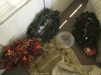 Photo showing two green Christmas wreaths, one decorated with beads and the other with a red bow, a Halloween pumpkin skull decoration to the right, orange-red fall-themed floral stems, a beige leaf patterned rug, and a clear candy cane-shaped glass serving dish in center.