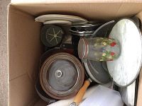 Box filled with assorted kitchen items including pots, pans, metal lids, a glass jar, and shelf paper, showing signs of age.