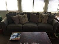 Wide front angle of the sage green cotton twill sofa with six throw pillows, showing sofa size and fabric texture in natural lighting.