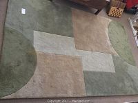 Large wool area rug with green, beige, and cream geometric pattern, showing wear and soiling from above.