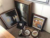 Seven pieces of wall art including two framed paintings with landscape scenes, framed movie poster titled 'The Hurricane', two decorative plates with scenic illustrations, framed photograph in black frame, and two small decorative wall plaques. Items rest on carpeted floor showing frame wear and age.