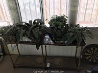 Four faux plants arranged on a woven and wooden console table under a window, showing all plants and planters in one view.