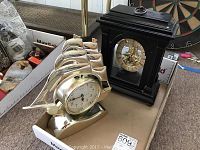 Two mantle clocks, one black wooden clock with visible gears and one gold plastic ship-shaped clock in a box.