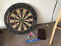 Dartboard leaning against wall with playing cards, pool chalk, and golf tees on floor in front of it.