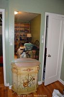 Vintage painted vanity with drawer, doors, and floral music motifs, minor chip visible on right side, against green wall beneath large rectangular mounted mirror reflecting bedroom.