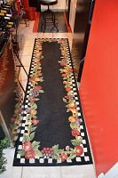 Kitchen area showing long Sonoma Fruit runner rug with black center and colorful fruit border on tiled floor, and black rubber mat with red border under stool.
